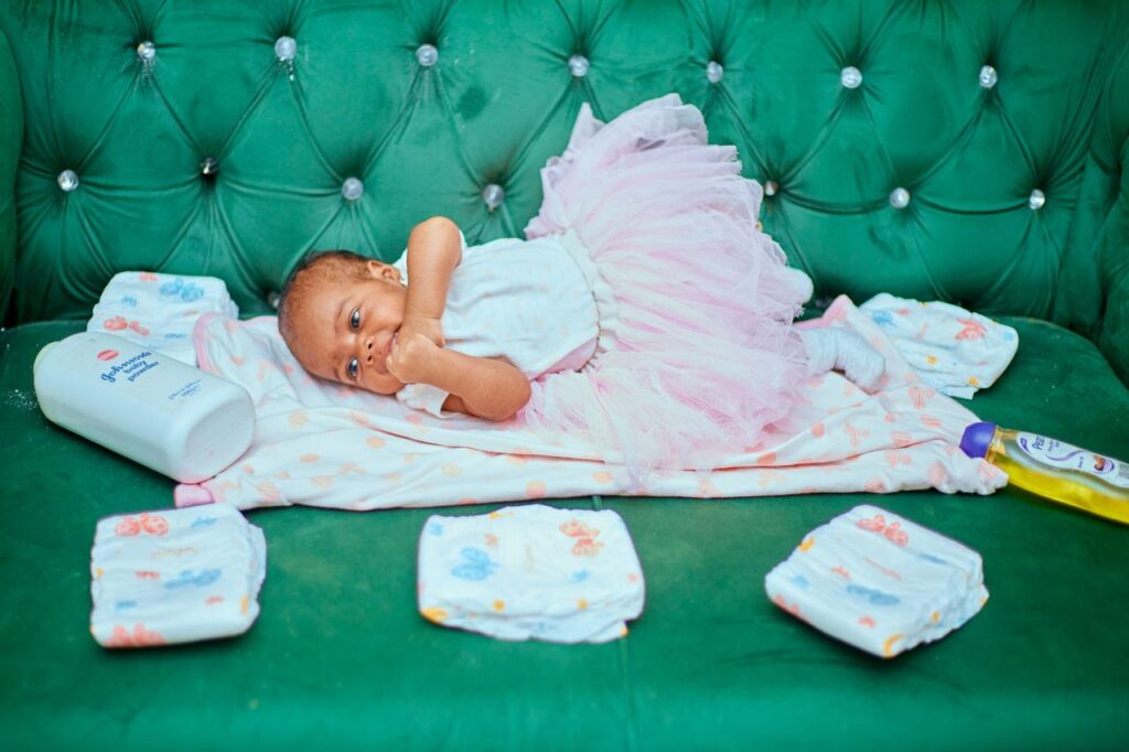 Cute baby lying on green couch with diapers and baby care products, wearing a pink dress.